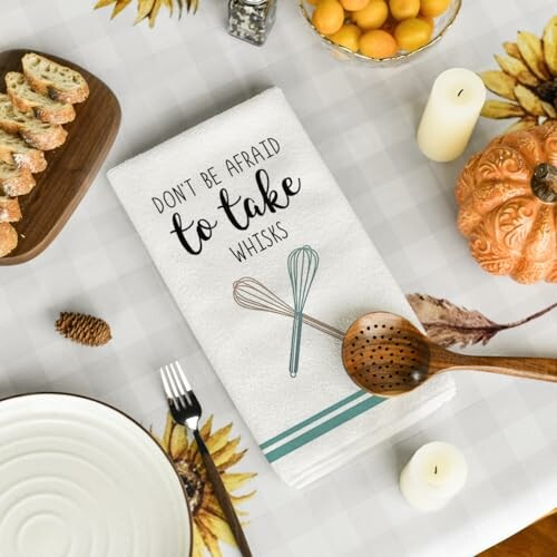 Festive table setting with a towel featuring 'Don't be afraid to take whisks' and kitchen items.